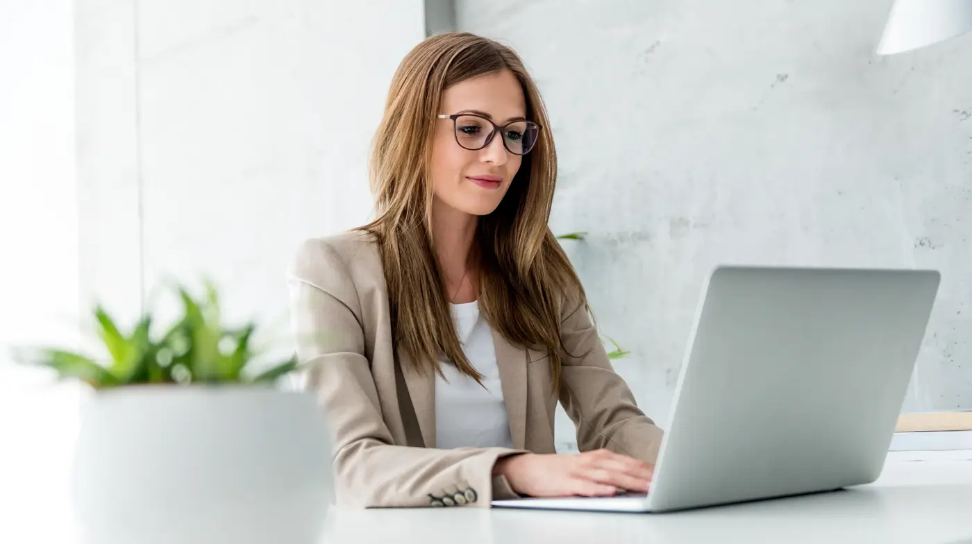 A woman wearing glasses is focused on her laptop, engaged in work at a desk.