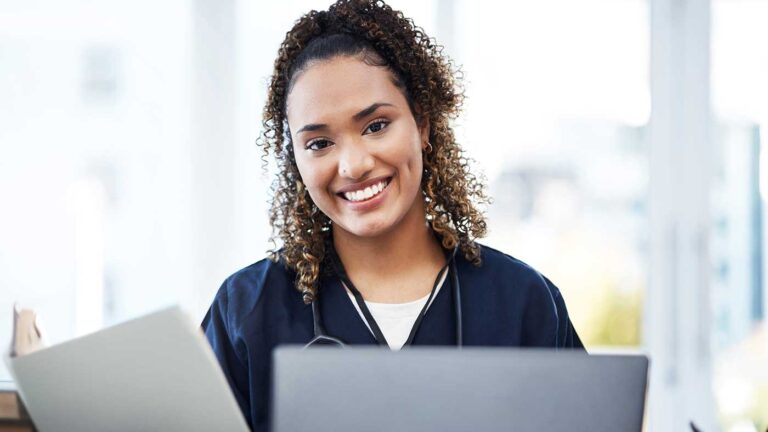 A smiling physician in a blue shirt sits at a desk, working on her laptop with a cheerful expression.